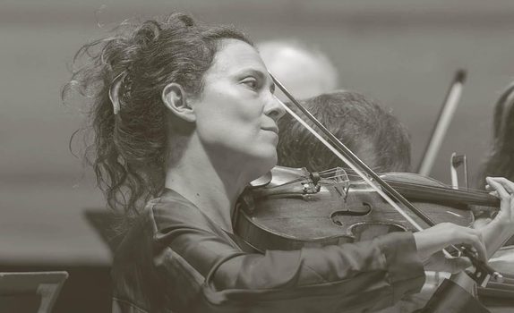 Lorenza Borrani plays the violin while the orchestra plays behind.