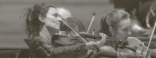 Lorenza Borrani plays the violin while the orchestra plays behind.