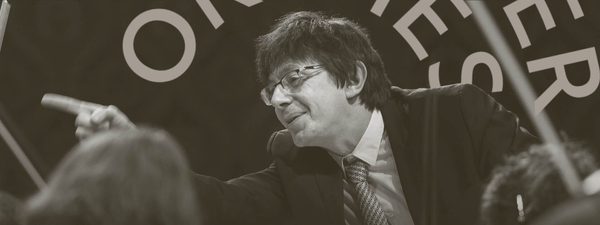 Vaclav Luks conducts. He points off to the right. He wears a patterened tie, glasses and a black jacket.