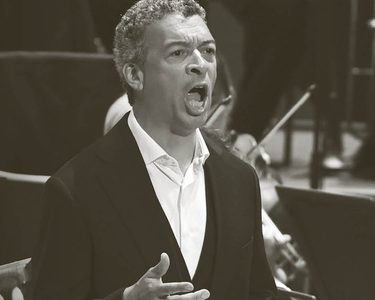 Roderick sings with the orchestra playing behind. He wears a white unbuttoned shirt and a black jacket.