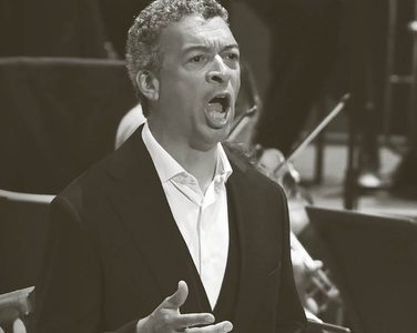 Roderick sings with the orchestra playing behind. He wears a white unbuttoned shirt and a black jacket.