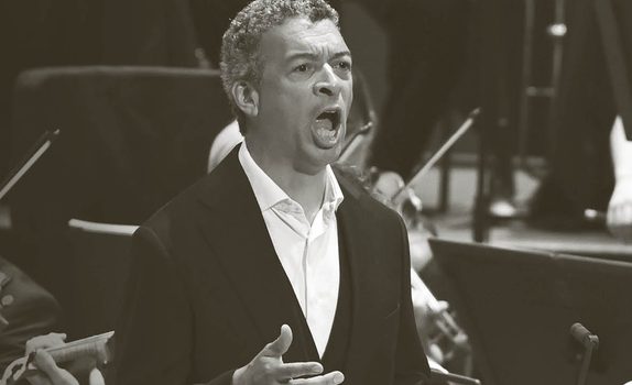 Roderick sings with the orchestra playing behind. He wears a white unbuttoned shirt and a black jacket.