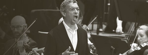 Roderick sings with the orchestra playing behind. He wears a white unbuttoned shirt and a black jacket.