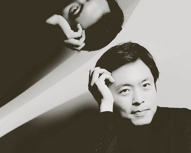 Sunwook rests his head on his hands. His image reflects above on the open piano case.