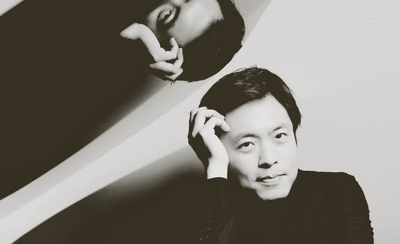 Sunwook rests his head on his hands. His image reflects above on the open piano case.