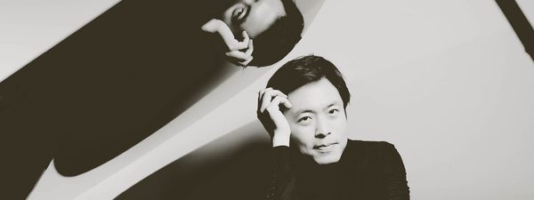 Sunwook rests his head on his hands. His image reflects above on the open piano case.