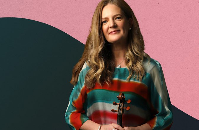Violinist Rachel Podger holds her instrument, smiling at the camera, against a light pink and dark green background.