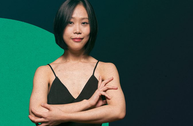 Pianist Yeol Eum Son standing with her arms crossed elegantly, lightly smiling at the camera, against a dark green background with a lighter green circle.