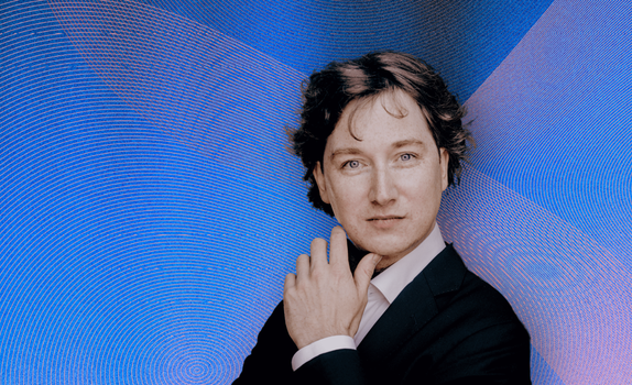 Maxim Emelyanychev looking into the camera, hand touching his jawline, against a gradient background of blue, pink, and orange waves.