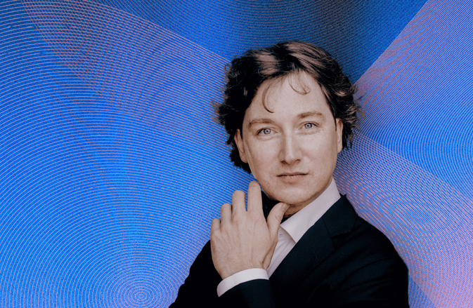 Maxim Emelyanychev looking into the camera, hand touching his jawline, against a gradient background of blue, pink, and orange waves.