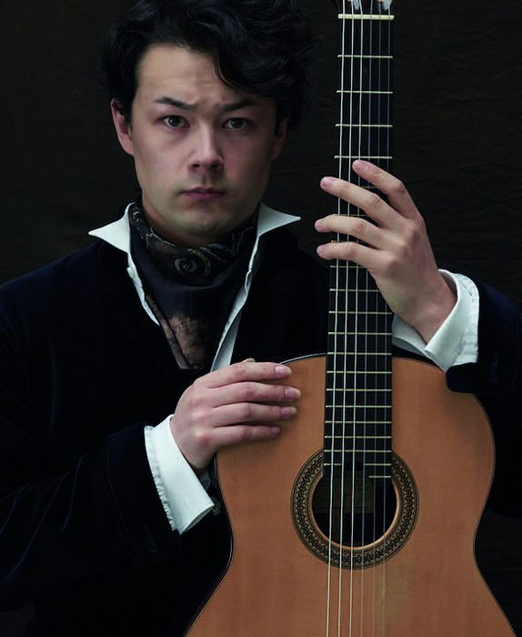 Sean shibe hold his guitar. He wears a white shirt with a high colour