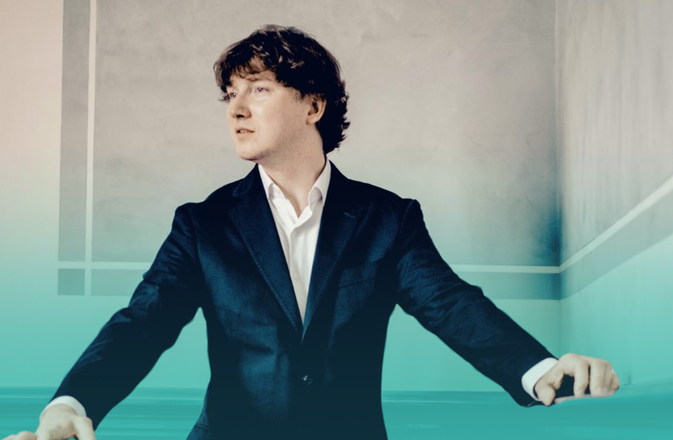 Maxim wears a suit and looks to the left of screen