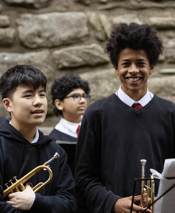 Two young teenage boys stand with trumpets in front of the Craigmillar Castle walls. One is looking to the music sheet, while the other looks into the camera and smiles.