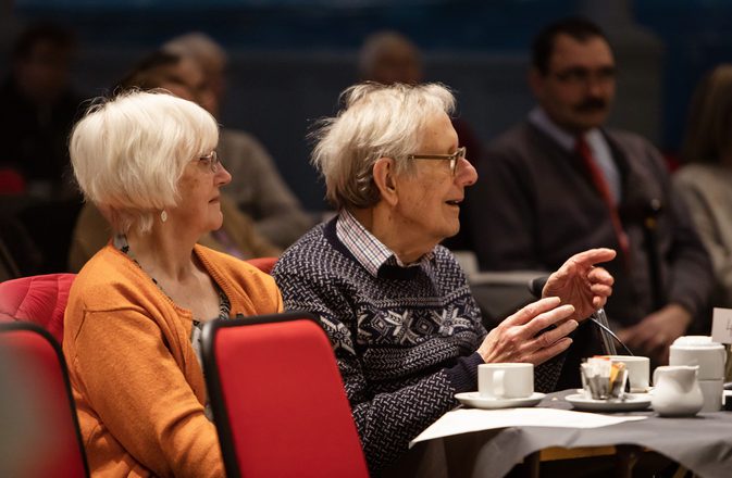 Two older people sit an smile with teas as they watch a performance.