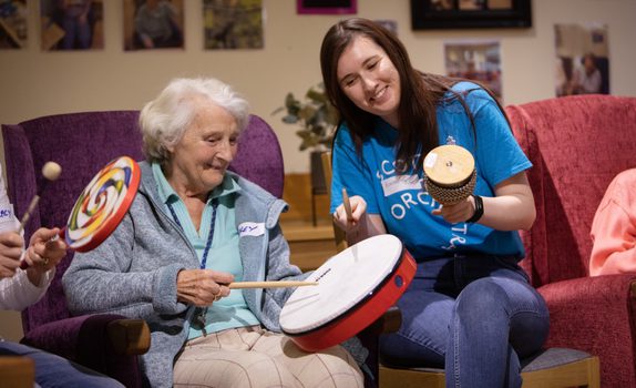 An SCO Creative Learning team member in a blue SCO tshirt holds a small bongo as an older lady smiles playing a handheld drum in the chair next to her.