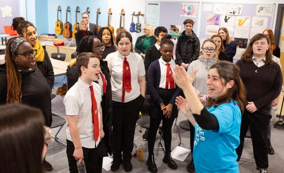 SCO Voices Schools Choir sing with red ties as leader Moira in a blue SCO t-shirt stands in front with her arm in the air.