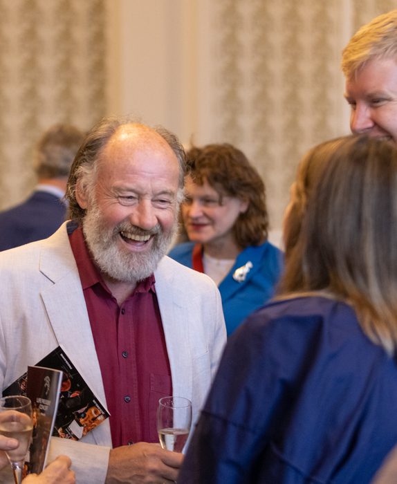An SCO donor smiles during a reception. He has a grey beard and wears an off white blazer with a maroon shirt.