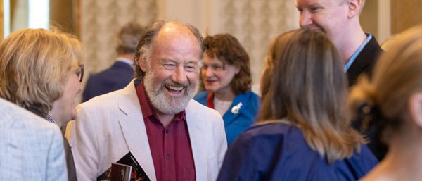 An SCO donor smiles during a reception. He has a grey beard and wears an off white blazer with a maroon shirt.