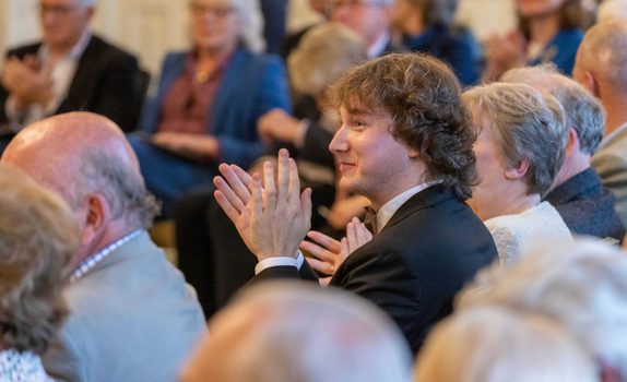 Maxim wears a black suit. He is clapping in an audience watching a performance.