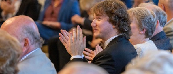 Maxim wears a black suit. He is clapping in an audience watching a performance.