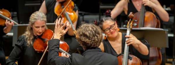 Back shot of Maixm conducting the orchestra. He wears a black jacket and raises his arm.