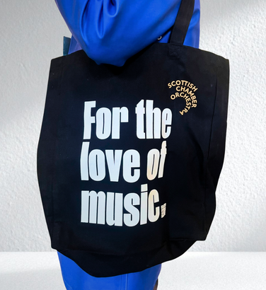 A person holds a black tote bag featuring the SCO logo in orange and For the Love of Music displayed in white text.