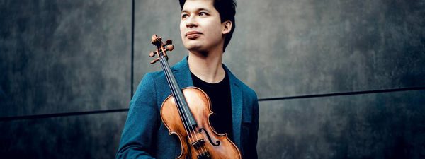A man stands against a black background made up of four tiles. He looks off to his right and is holding a violin at its base. Its front faces the camera.