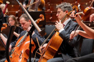 Two men play the cello in an orchestra, facing left
