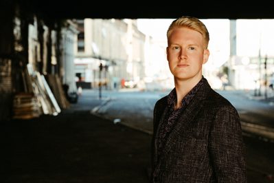 A man in a blazer and shirt looks towards the camera. Behind the composer is a city backdrop.