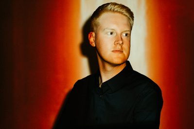 Against a red background, a man in a dark jacket and shirt looks out of frame. His face is illuminated by a light from above.