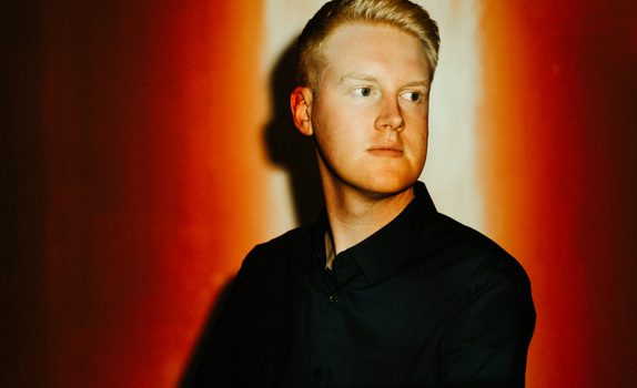 Against a red background, a man in a dark jacket and shirt looks out of frame. His face is illuminated by a light from above.