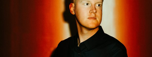 Against a red background, a man in a dark jacket and shirt looks out of frame. His face is illuminated by a light from above.