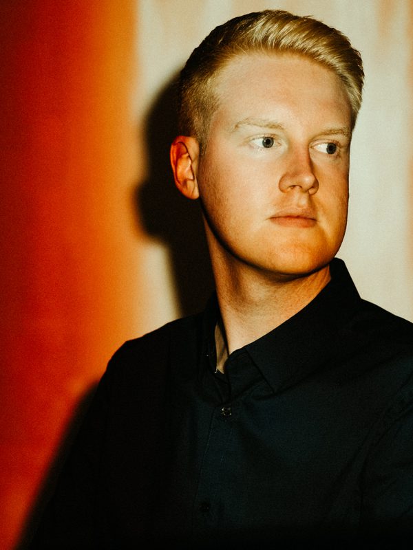 Against a red background, a man in a dark jacket and shirt looks out of frame. His face is illuminated by a light from above.