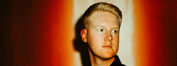 Against a red background, a man in a dark jacket and shirt looks out of frame. His face is illuminated by a light from above.