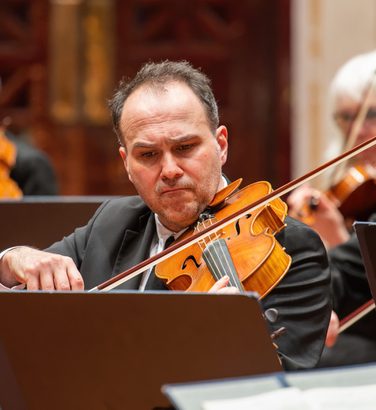 Max Mandel plays the viola, concentrating hard on the sheet music. He wears a black suit and tie.
