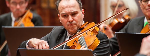 Max Mandel plays the viola, concentrating hard on the sheet music. He wears a black suit and tie.