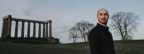 A man stands in profile (facing the camera) on a hillside, framed by two trees, with a series of stone columns from an unfinished temple on the far left