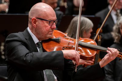 Steve King plays viola. He wears a black suit and tie and black glasses.