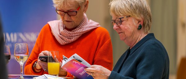 Two audience members read a programme. They are older women, one wears a red jumper and scarf, the other has a blue jumper on. Both wear glasses.
