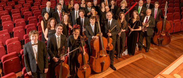The Orchestra stand in a hall staring up to the camera. Maxim join to the left side.