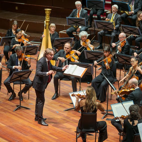Maxim conducts the orchestra from the Usher Hall. He wears a black suit.