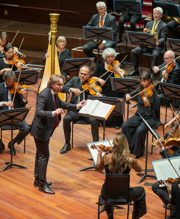 Maxim conducts the orchestra from the Usher Hall. He wears a black suit.