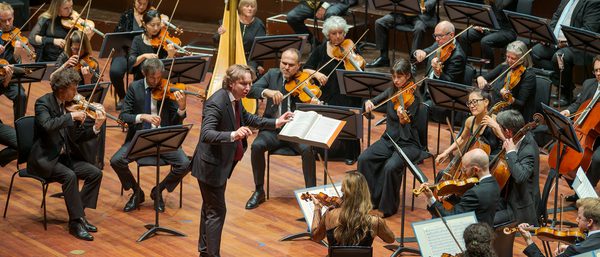 Maxim conducts the orchestra from the Usher Hall. He wears a black suit.