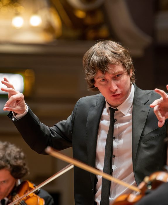 Maxim conducts the orchestra. String players are in shot.