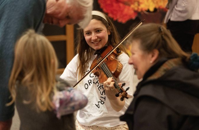 An SCO player with the Creative Learning team, demonstrates the violin to a child, as her grandparents watch.