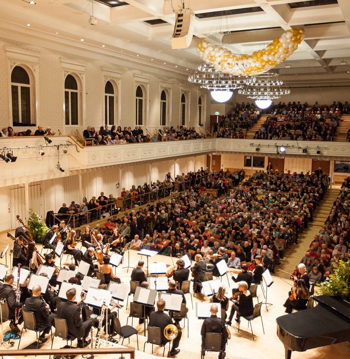 The orchestra take their seats on stage in front of the audience of a sold out concert.